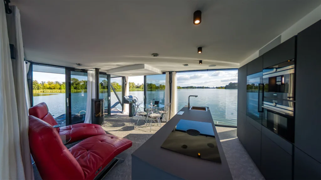 Offener Wohnbereich mit Designküche, roten Relaxsesseln und Panoramafenstern zum See