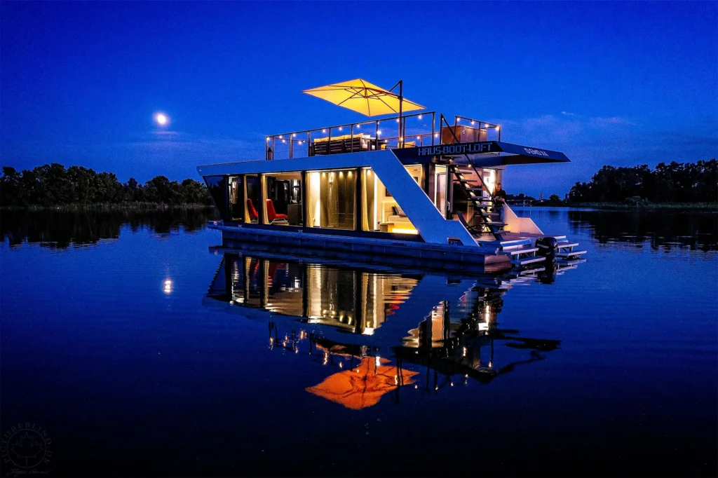 Beleuchtetes Hausboot bei Nacht auf dem See mit Mond und Spiegelung im Wasser