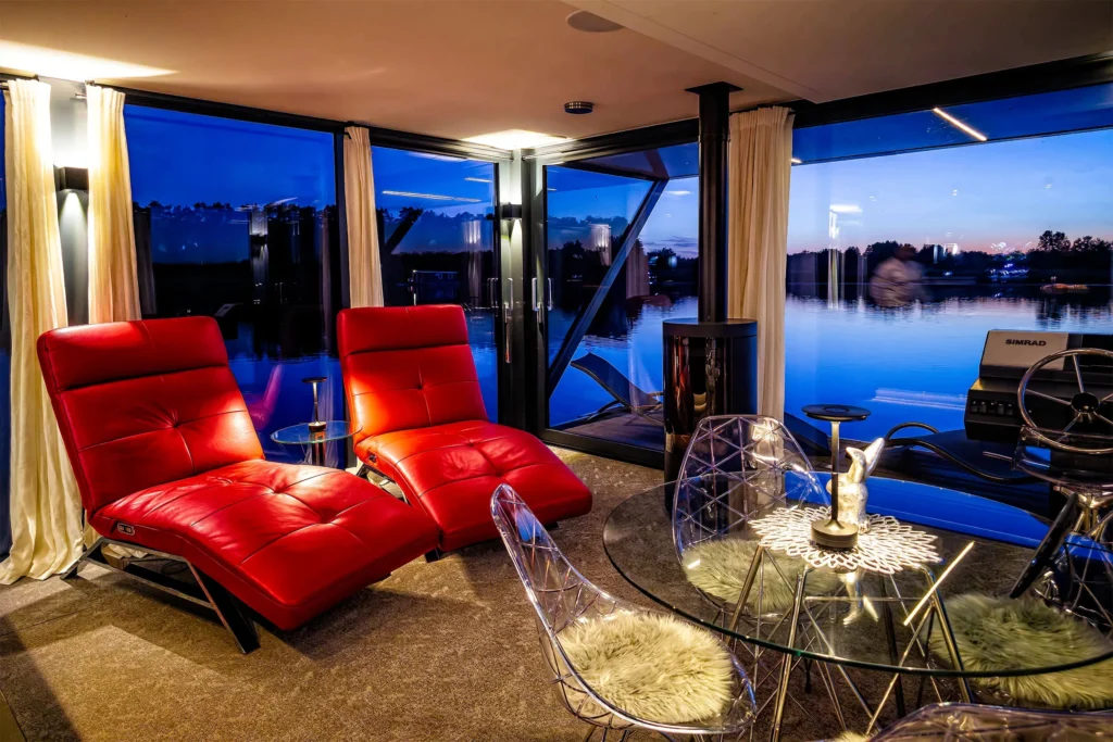 Wohnbereich am Abend mit zwei roten Relaxliegen, Glastisch, Kamin und Seeblick durch die Glasfront