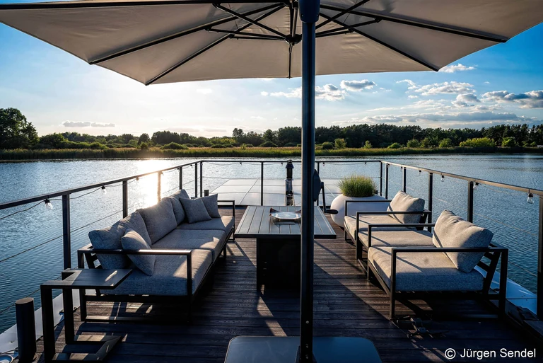 Dachterrasse mit Lounge-Sitzgruppe und Sonnenschirm im Gegenlicht über dem See