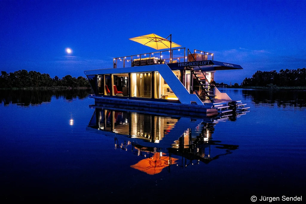 Beleuchtetes Hausboot bei Nacht auf dem See mit Mond und Spiegelung im Wasser