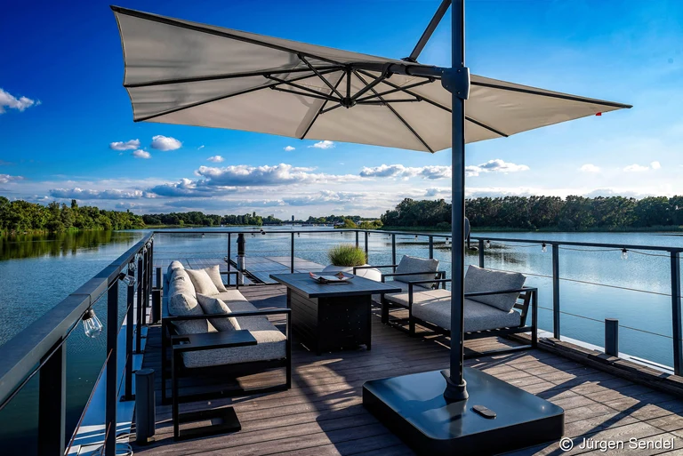 Heckterrasse des Hausboots mit Lounge-Sofa, Sesseln und Sonnenschirm direkt über der Havel an einem Sommertag
