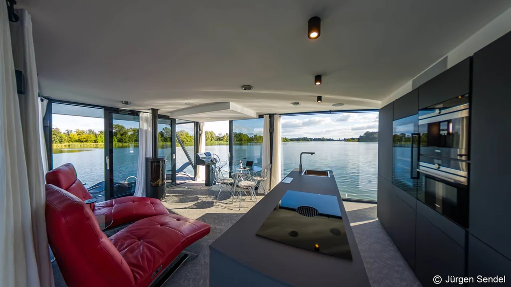 Offener Wohnbereich mit Designküche, roten Relaxsesseln und Panoramafenstern zum See